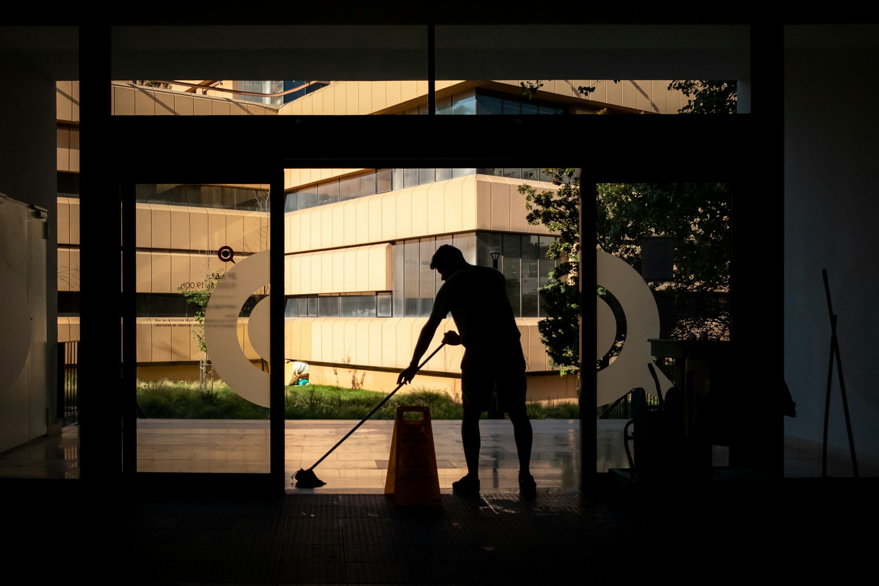Silhouette of a cleaner mopping a glass lobby at dusk